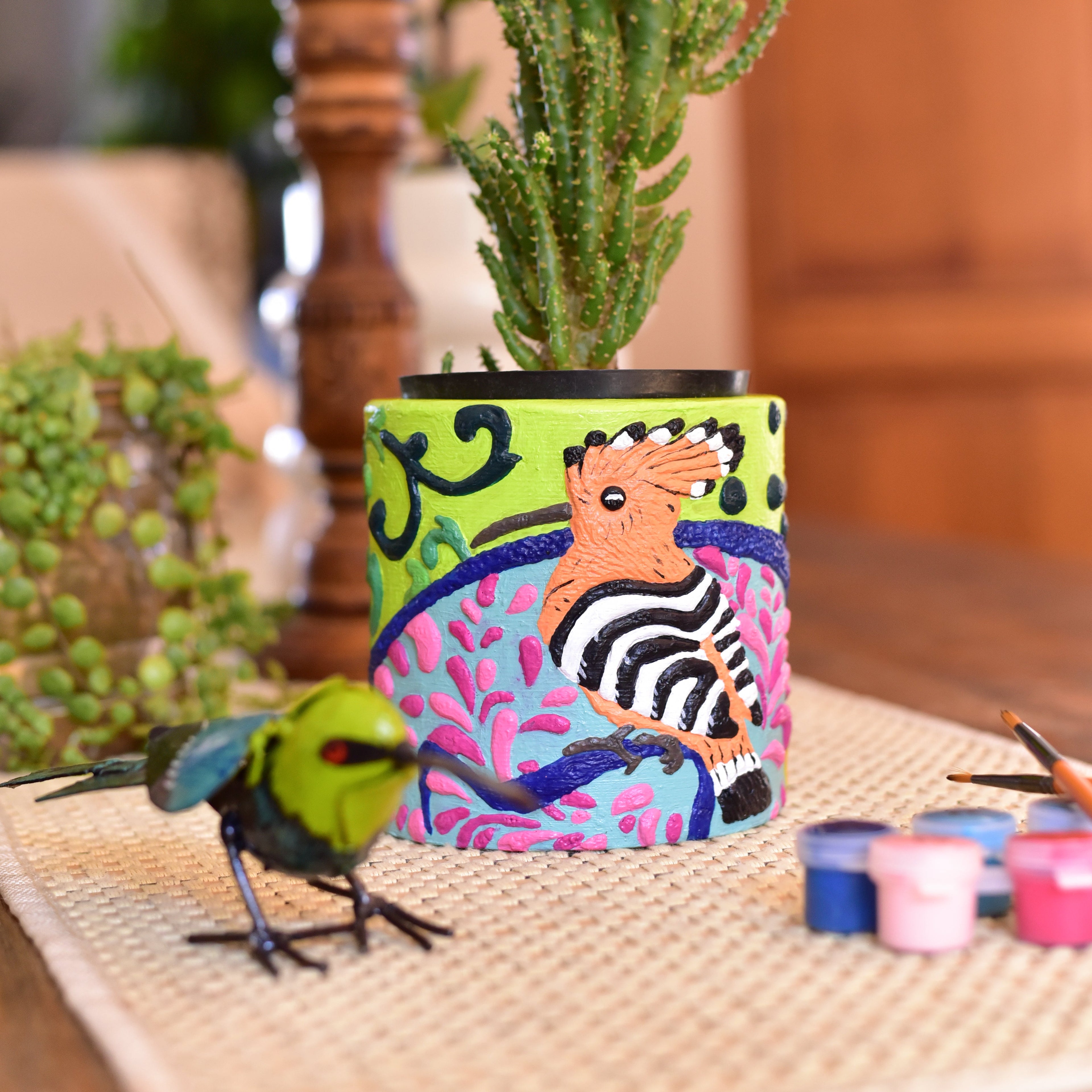A colorful hand-painted paint-by-numbers planter with a hoopoe and pink  design, holding a green succulent.