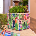 A square paint-by-numbers planter decorated with a colorful desert floral and meercat illustration, filled with green succulents, displayed on a wooden table with paint supplies nearby.