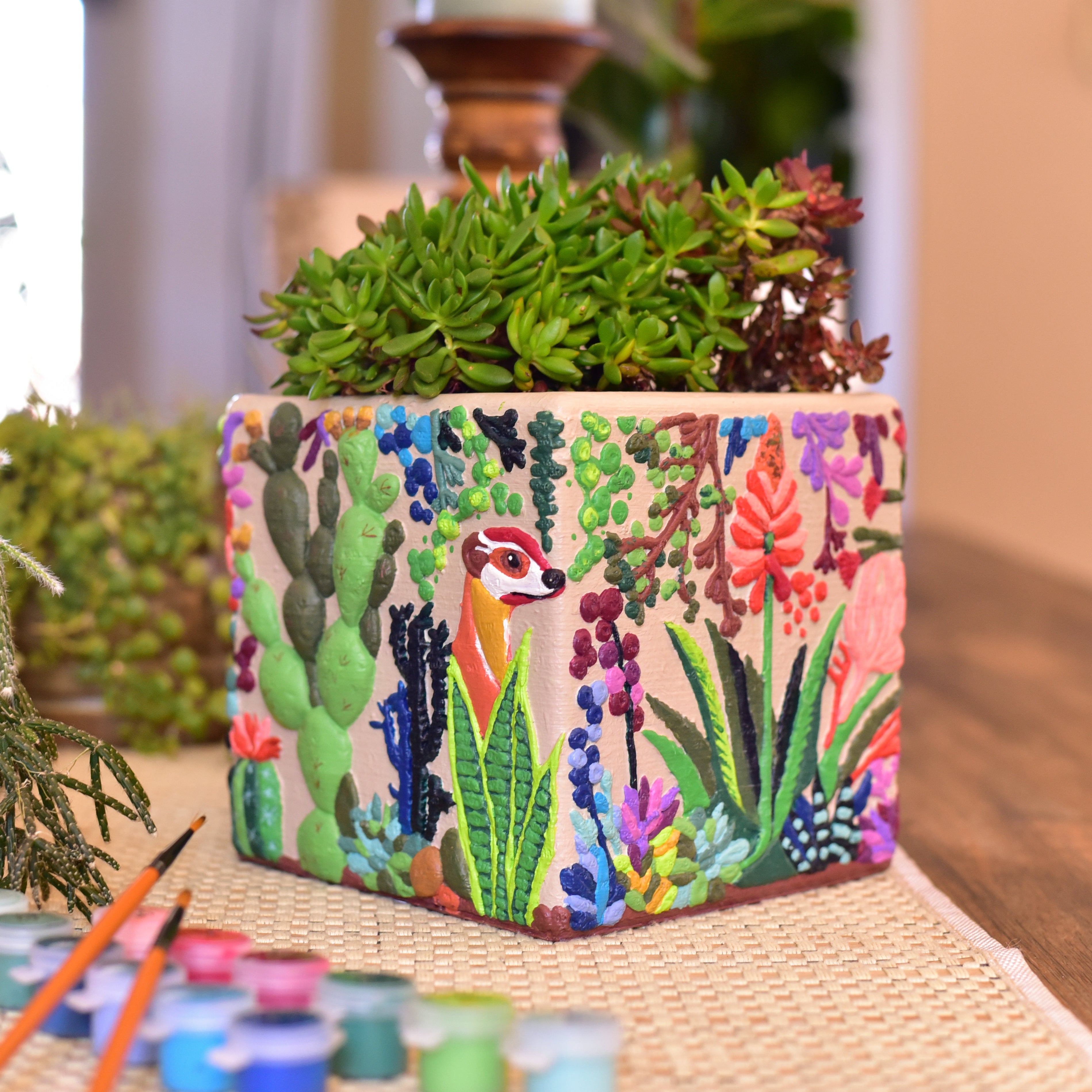 A square paint-by-numbers planter decorated with a colorful desert floral and meercat illustration, filled with green succulents, displayed on a wooden table with paint supplies nearby.