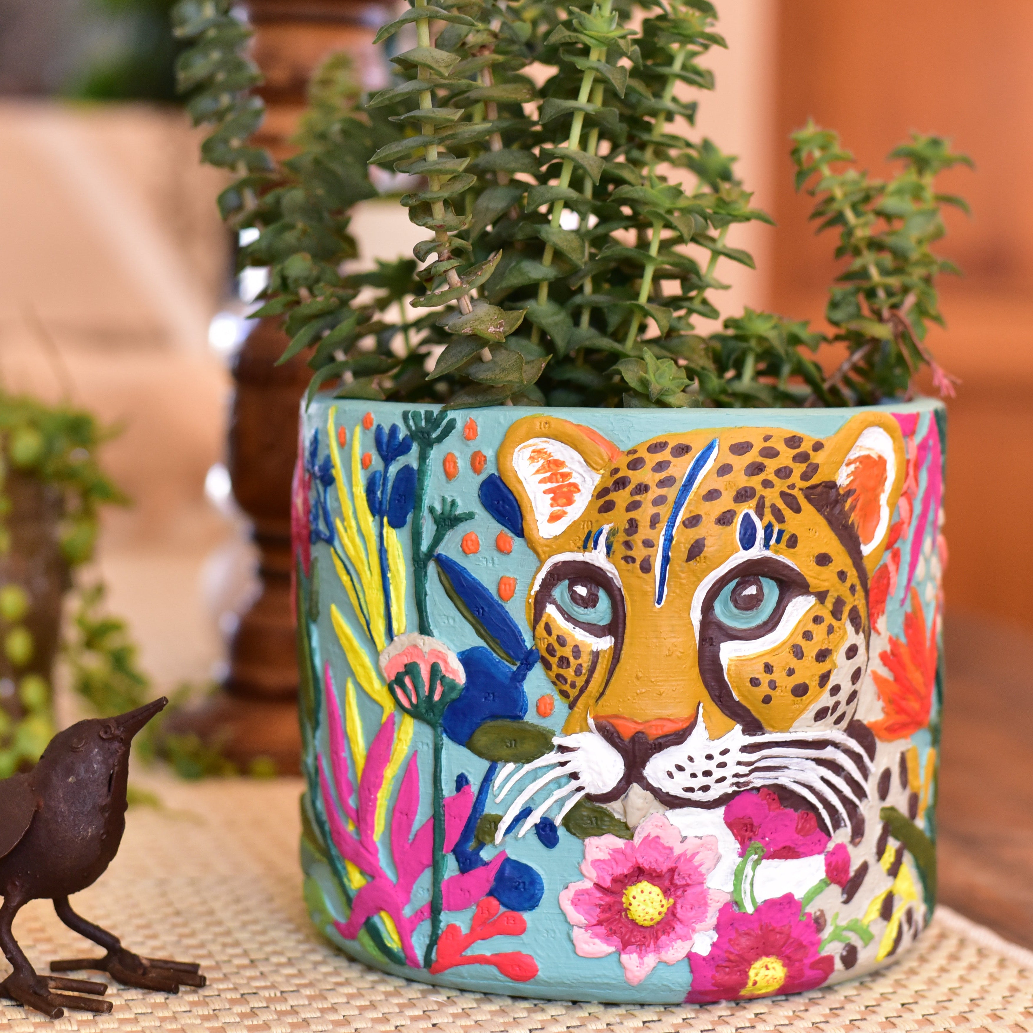 A colorful paint-by-numbers planter featuring a stylized cheetah face and  floral motifs, filled with green succulent stems.